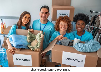 Small Mixed Race Group Of People Working In Charitable Foundation. Male And Female Teenage Food Bank Volunteers Sort Canned Food Items In Cardboard Boxes. Happy Volunteer In Community Food Bank