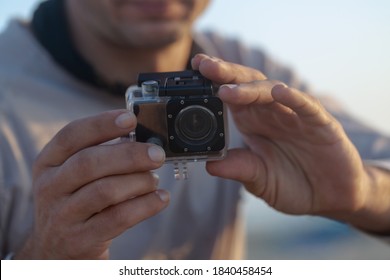 Small Modern Action Camera In Waterproof Housing Filming In Shallow Water On Beach, Man Holding Recording Equipment Sitting Outdoors. Selective Focus