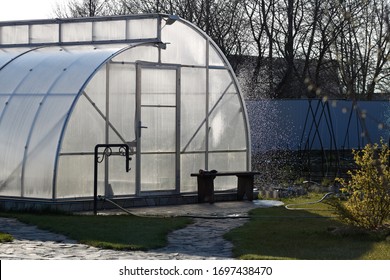 A small modern greenhouse made of aluminum frame and transparent polycarbonate is built in a private yard for growing organic vegetables and greens. Honeysuckle bush is out of focus. Water splashes in