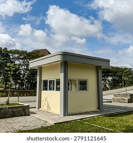 A small modern security guard post or booth building featuring yellow painted walls, grey concrete flat roof with extended canopy, and aluminium sliding windows. The structure stands on a paved courtyard surrounded by neatly landscaped grounds with young trees, stone planters, and green grass. A dramatic blue sky with large white cumulus clouds provides a vivid backdrop, while lush tropical trees fill the middle ground. Photographed in Magelang, Central Java, Indonesia, this image is suitable for architecture, security, urban infrastructure, and institutional facility themed content.