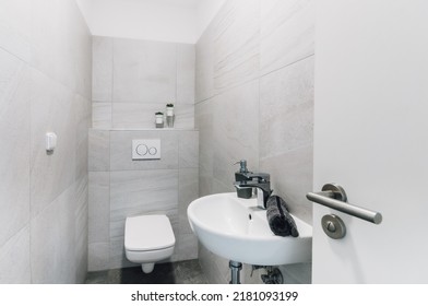 Small modern toilet with sink, towel and soap dispenser. On the surrounding ones, we can find a switch, a flush button and the part of the door with a handle and lock visible to the right. - Shutterstock ID 2181093199