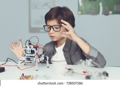 A small nerd in glasses is holding a robot. A boy in a gray shirt and glasses is sitting at a table in front of him whose robot the boy made. He is very satisfied with his work