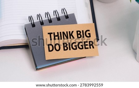 Small notepad rests on white surface. Bold text reads: Think Big Do Bigger. Open notebook background suggests planning or ambition. Minimalist scene evokes motivation and progress