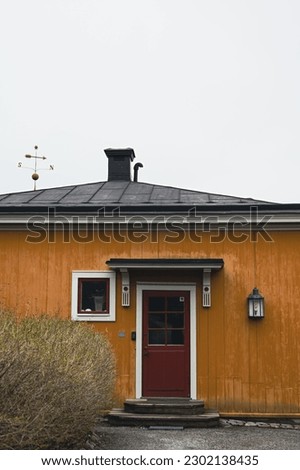 A small, old, orange, Scandinavian house from the countryside of Sweden, with an outside lamp, a red door and a single window, with a bush outside and a compass on the roof, on a foggy day