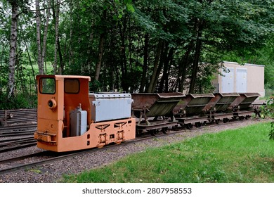 A small old orange train engine with a row of V-shaped dump metal carts parked on a railway line surrounded by green grass.