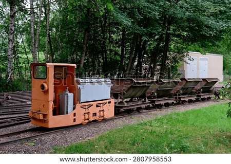 A small old orange train engine with a row of V-shaped dump metal carts parked on a railway line surrounded by green grass.