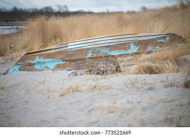 A small old row boat on the beach with a broken lobster trap in the sand. The boat's blue paint is worn and peeling from the sides. The boat is laying upside down in the sand and dune grass.