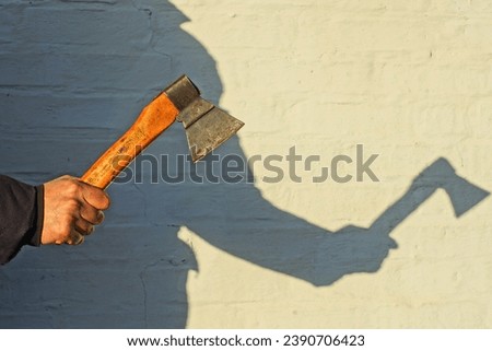 small old sharp dangerous iron industrial rusty with a wooden handle iron hatchet in hand near a white wall on the street with the shadow of a man's silhouette in the sun