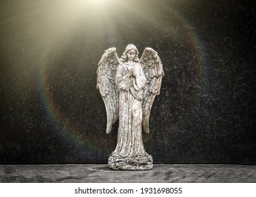 Small old weathered stone weeping angel with wings ornament standing in sorrow on black granite grave headstone in peaceful churchyard with heavenly sunlight golden rays shining in