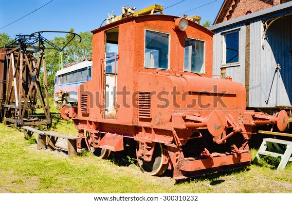 Small orange abandoned train wreck with\
some rust. Train car and other junk in background. Door is missing\
and no person is visible. Green grass on\
ground.