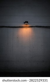Small orange wall light illuminating a weathered gray exterior wall with exposed electrical cables, creating a minimal urban scene with copy space