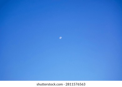 A small, pale crescent moon is visible in the middle of a vast, bright blue sky. The image conveys a sense of peace, minimalism, and daytime lunar visibility.