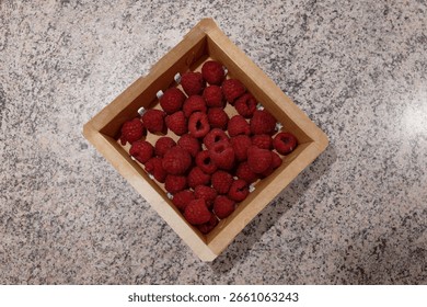 A small paper container filled with fresh red raspberries sits on a dark gray graphite table surface, showcasing a simple natural snack.