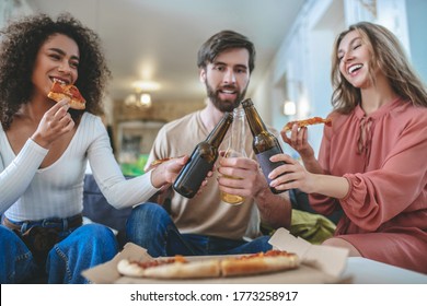 Small Party. Three Young Adult Cheerful Friends With Slices Of Pizza And Bottles Touching One Another, Sitting At Home Having Rest