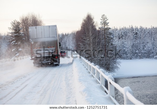 Small passenger car and lorry\
driving on bridge across river on snowy road at winter\
season