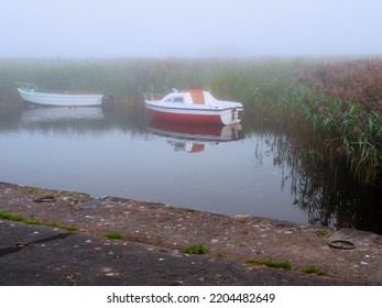 Small Pier By Lake With Tall Green Grass And Two Small Fishing Boats In Water. Fog In The Background. Calm And Peaceful Nature Scene. Nobody.