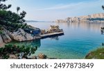 Small pier extends from rocky outcrop into calm reflective Adriatic sea at sunrise near Baska, Krk Otok, Primorje-Gorski Kotar, Croatia, Europe. Mountain silhouette in morning mist. Ethereal glow