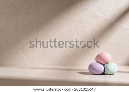A small pile of three speckled Easter eggs in soft pastel colors rests on a textured surface, illuminated by gentle light and shadow patterns