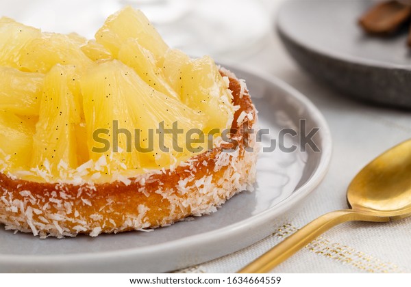Small pineapple tart or tartlet with glazed\
pineapple fruit pieces and grated coconut by a golden spoon, over a\
white and gold table\
cloth.