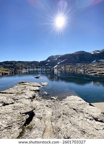 Small pond on Trolltunga path, mirroring rock formations, green moss clinging to stones. Clear sky, gleaming sun, calm water, vibrant reflections, peaceful Norwegian beauty.