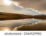 A small pool of water on rural farming fields reflecting the snow covered southern alps and a n amazing cloudscape
