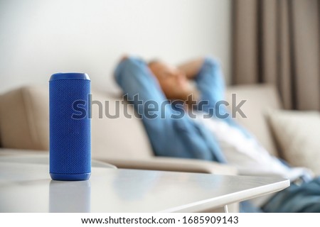 Small portable wireless speaker assistant on table with man lounge on sofa listening to music. Young relaxed guy enjoys mini bluetooth stereo gadget for sound digital assistance at smart home concept.