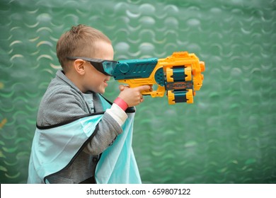 Small Preschooler Boy With Blaster Prepare For Attack And Play With Friends In Protective Glasses. Excited Child With Darts Toy Gun On The Play Field