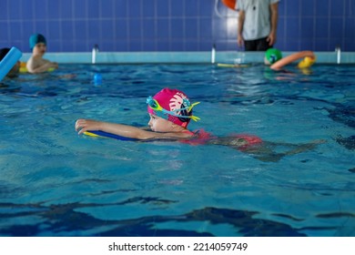 Small Preschooler Child Learns To Swim With Board In Pool. Swimming Lesson. Active Child Plays In Water. Kid In Mask And Goggles.