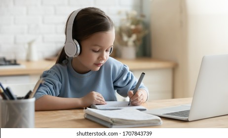 Small preschooler girl in headphones sit at desk study online on laptop, smart little kid wear earphones handwrite in notebook learning using internet lessons on quarantine, homeschooling concept