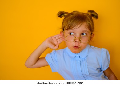 A small pretty girl on an orange background in a blue shirt and gold-rimmed glasses, listening carefully with her hand to her ear. Curious, surprised child