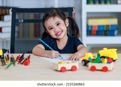 Small Pretty Pigtails Hair Preschooler Kindergarten Happy Girl Sit On Chair Drawing With Colored Pencils On Paper On Table Full Of Plastic Truck Toys In Living Room At Home In Front Bookshelf.