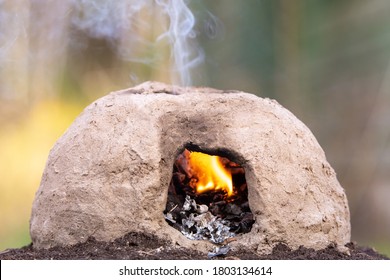 A Small, Primitive Oven Made From Mud. A Fire Burns Inside And Smoke Rises From A Hole In The Top.