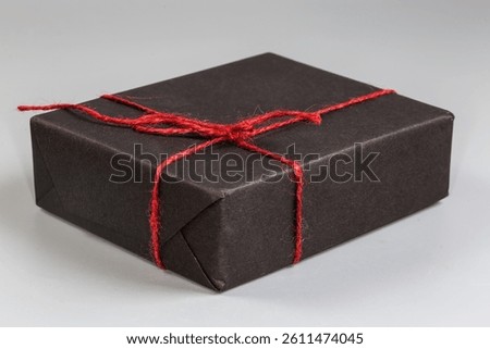 Small rectangular flat packing box wrapped in black wrapping paper and criss-cross tied with red rope, front view on a gray background