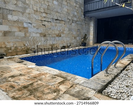 A small, rectangular swimming pool with bright blue tiles, silver handrails, and surrounding stone. A light-colored stone wall is behind the pool