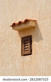 A small rectangular window with closed brown wooden shutters on a textured beige plaster facade featuring a small tiled roof canopy