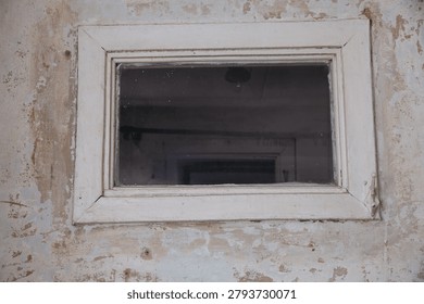 A small, rectangular window with a white frame set in a weathered wall. The glass is slightly dirty, revealing a dimly lit interior behind it.