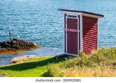 A small red building, outhouse, on the edge of a rocky cliff with blue ocean and islands in the background.  The outhouse has a closed sign across its door. The sky is blue with some white clouds. 