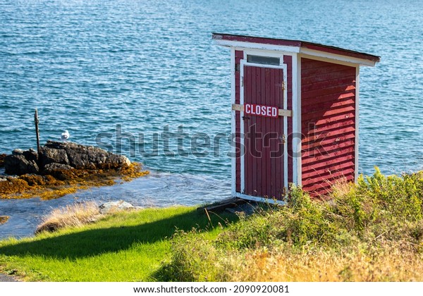 A small red building, outhouse, on the edge of a\
rocky cliff with blue ocean and islands in the background.  The\
outhouse has a closed sign across its door. The sky is blue with\
some white clouds. 