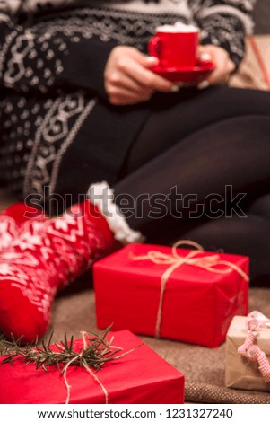A small red cup with hot chocolate and marshmallows in the hands of a woman in red warm socks sitting among packed Christmas gifts