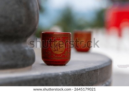 A small red cup is placed in front of the altar of worship, the cup is decorated with intricate traditional Chinese motifs in gold, a similar cup is visible in the background with a blurred effect