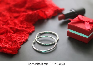 Small red giftbox, red lipstick, silver earrings and lace underpants. Valentine's Day objects on dark background. Selective focus.