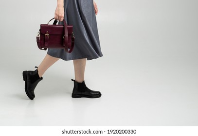 Small Red Leather Bag In A Woman's Hand On A White Background. Shoulder Handbag. Woman In A White Shirt And Grey Skirt And With A Red Handbag. Style, Retro, Fashion, Vintage And Elegance.