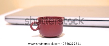 Small red maroon mug cup on white table beside a sleek laptop with dimming soft red light, web banner laptop soft color