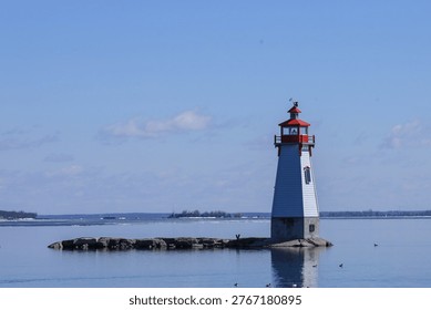 Small red and white lighthouse at Jackson's Point on Lake Simcoe, Ontario, Canada, set on a rocky shoreline with visible layers of water, distant land, and soft white clouds.  Horizontal composition with subtle reflection and a calm, scenic lakeside atmosphere. 