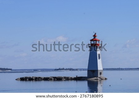 Small red and white lighthouse at Jackson's Point on Lake Simcoe, Ontario, Canada, set on a rocky shoreline with visible layers of water, distant land, and soft white clouds.  Horizontal composition with subtle reflection and a calm, scenic lakeside atmosphere. 