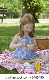 Small Red-haired Girl Eating A Cup Of Applesauce Outdoors In A Park - Picnic Setting.