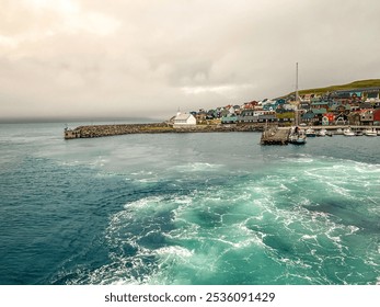 Small Remote Island of Nólsoy as Viewed from the North Atlantic Ocean. Wake and waves from an arriving ferry boat. Boats moored in the harbor. Located within the archipelago of the Faroe Islands. - Powered by Shutterstock