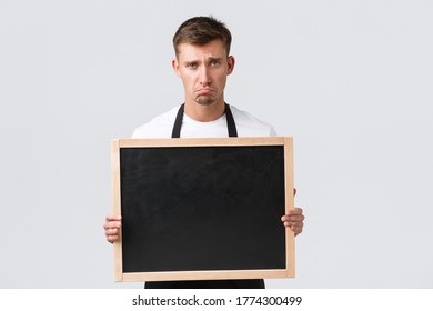 Small Retail Business Owners, Cafe And Restaurant Employees Concept. Disappointed And Sad Salesman In Black Apron Pouting And Looking Unhappy, Showing Blank Board For Client Info