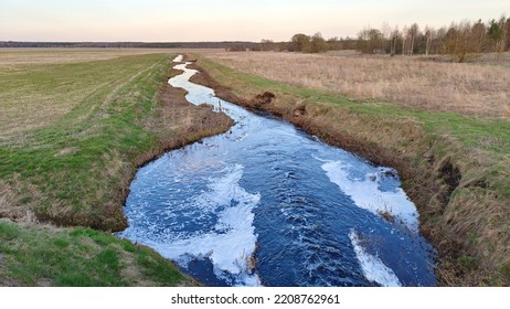 A Small River Runs Through Grassy Meadows To The Forest On The Horizon. After Passing The Dam, The Water In The River Bubbles And Foams. The Spring Evening Twilight Is Setting