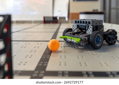 A small robot navigates a table while targeting an orange ball in a tech workshop.